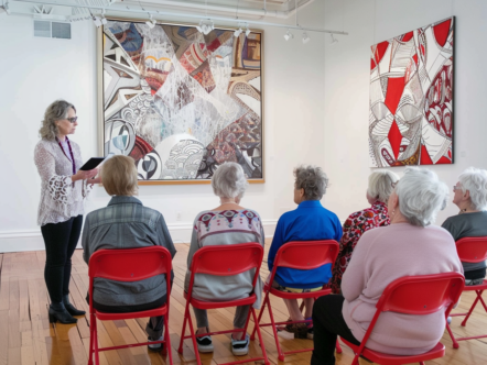 photorealistic group of elderly people sitting on red folding chairs in an art gallery, listening to the hostess explaining about one painting hanging from above them. The artwork is of abstract patterns and shapes, the room has white walls with wood floors, there's another large canvas hanging behind it, the setting feels calm and relaxing --no blur --ar 4:3