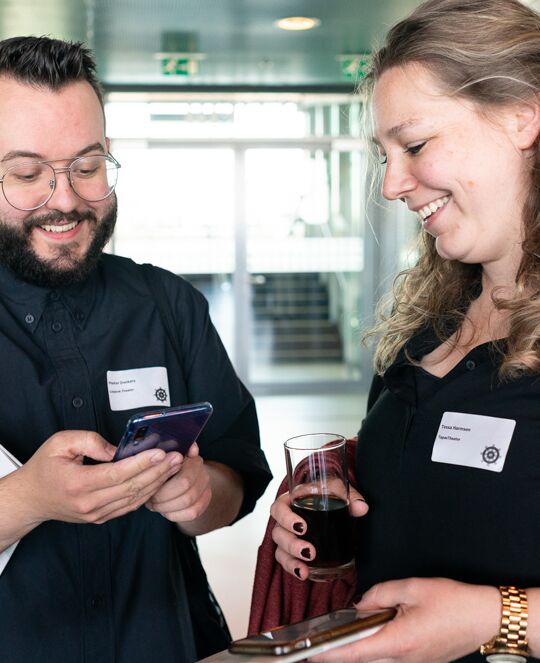 Word lid van Cultuurmarketing. Twee mensen kijken lachend naar een telefoon. Eén houdt een drankje vast.